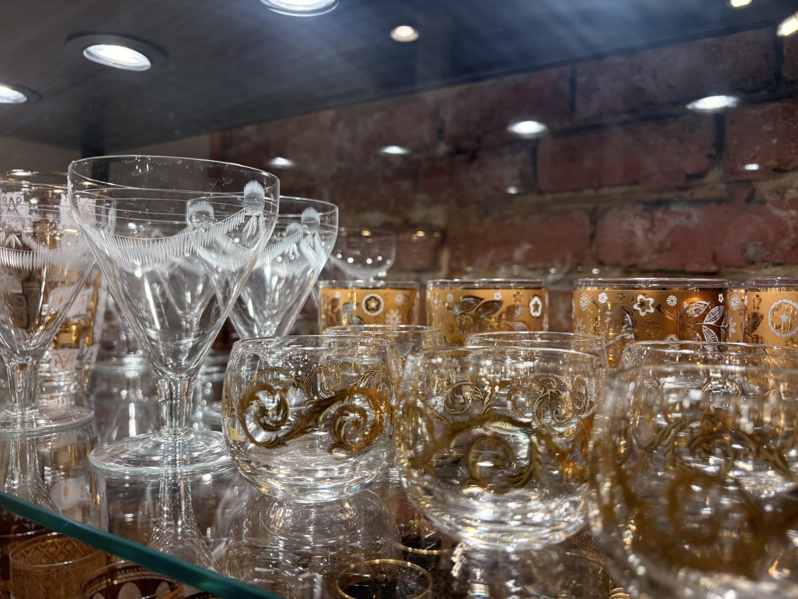 Gold-patterned mid-century rocks glasses and cut-crystal coupes on a glass shelf against exposed brick